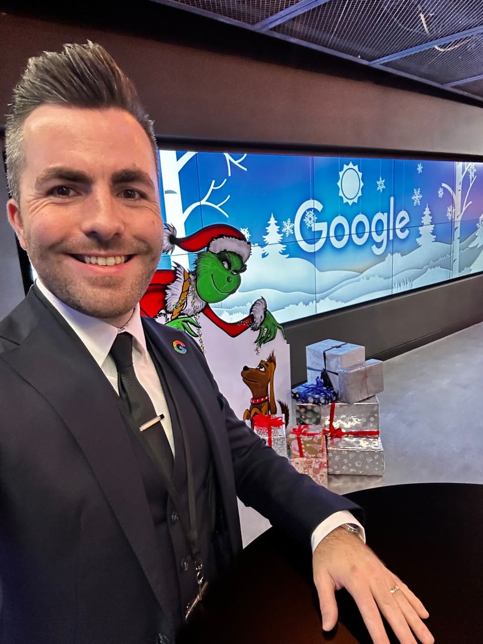 Jamie smiles in front of a wall of screens with a snowy Google logo, a Grinch cut-out and a pile of wrapped gifts