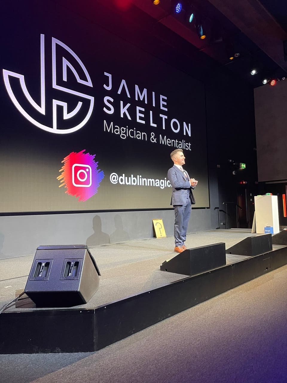 Jamie on a big stage in a three-piece suit, his name on the screen behind him, a small Google sign on a plinth