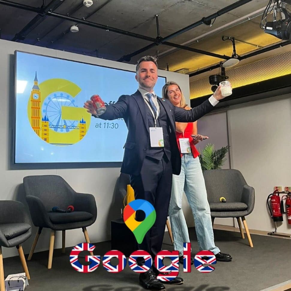 Jamie on a small stage at Google in London holds up a can and a cup, a volunteer behind him, a London-landmarks G on the screen