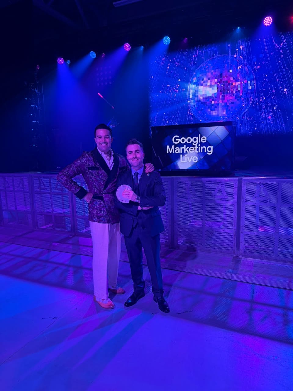 Jamie holds a fan of cards beside a performer in a brocade jacket, in front of a stage screen that reads Google Marketing Live