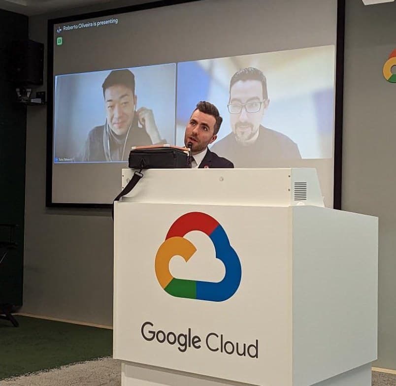 Jamie at a Google Cloud lectern, on a big screen beside two colleagues on a video call
