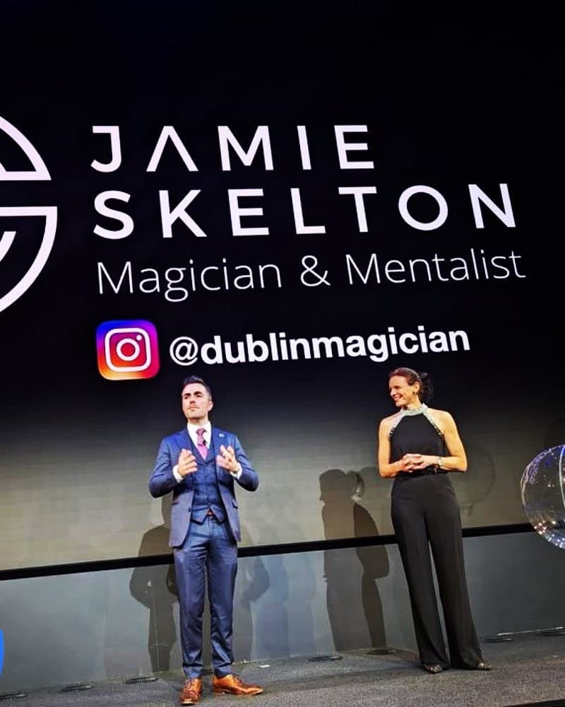 Jamie talks to the room from a dark stage beside the host in a black jumpsuit, his name on the big screen behind them