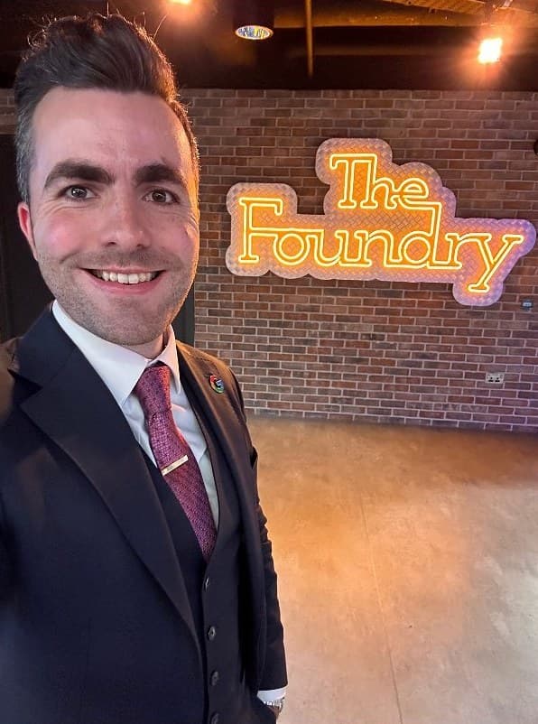Jamie smiles in a three-piece suit beside a neon sign that reads The Foundry