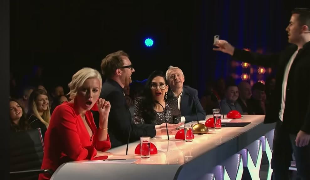 Jamie holds up a small object beside the Ireland's Got Talent judges' desk as the four judges watch
