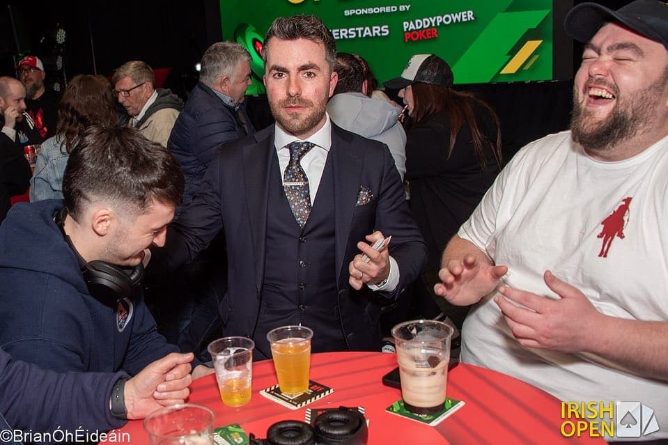 Jamie holds a card at a red poker table while a player in white roars laughing beside him