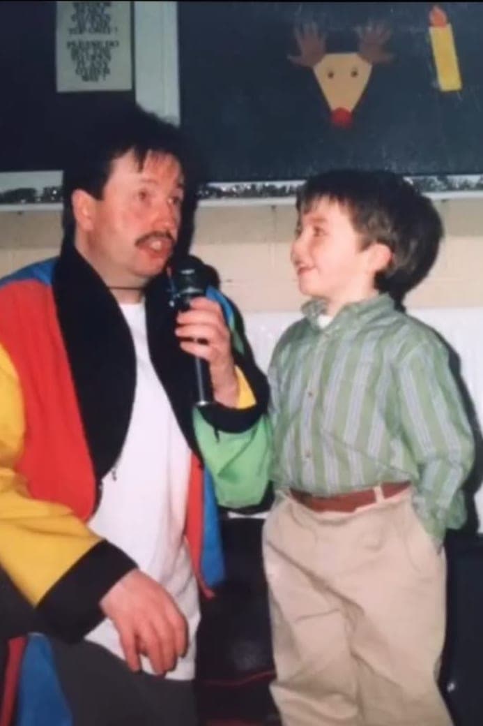 A young Jamie stands on stage beside a magician who holds a microphone