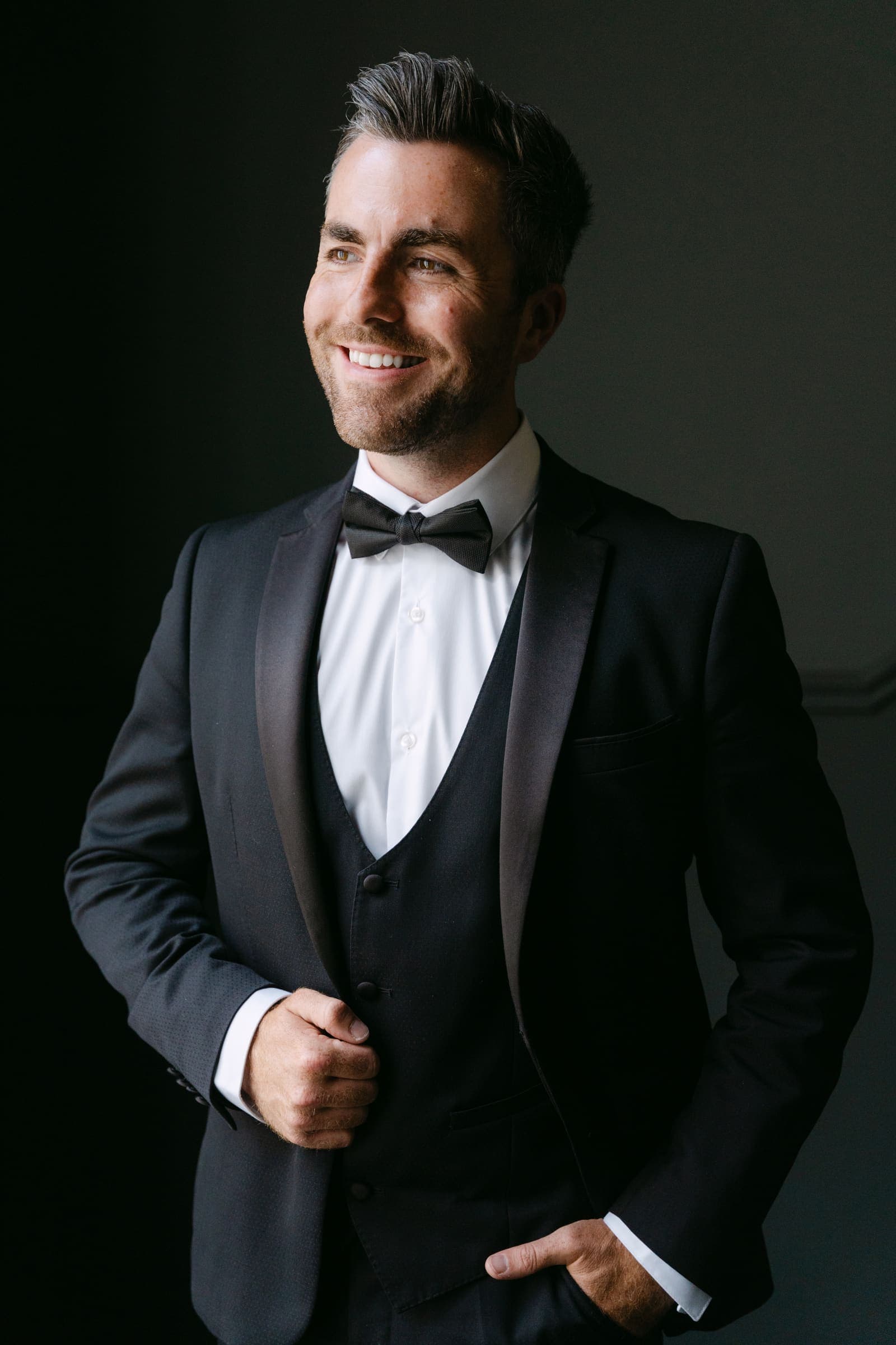 Headshot of Jamie Skelton smiling in a black tuxedo and bow tie against a dark wall