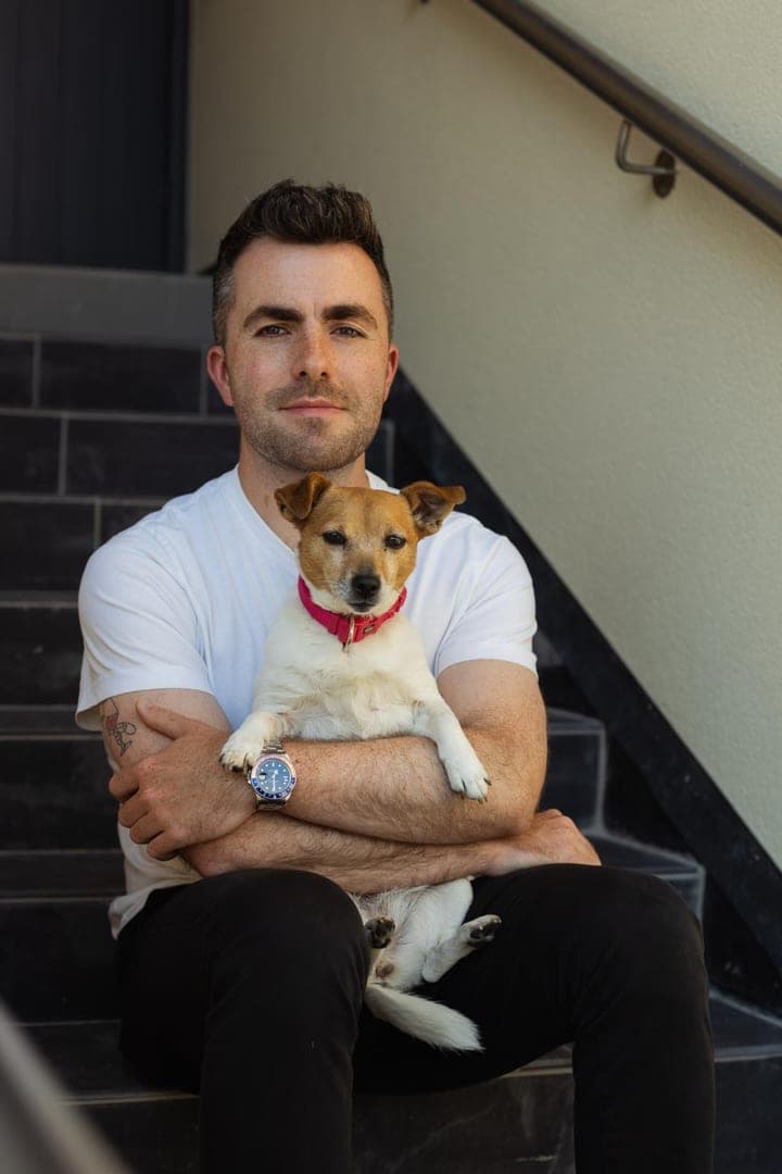 Jamie sits on the stairs at home and holds his rescue dog Piper, a small white and tan terrier