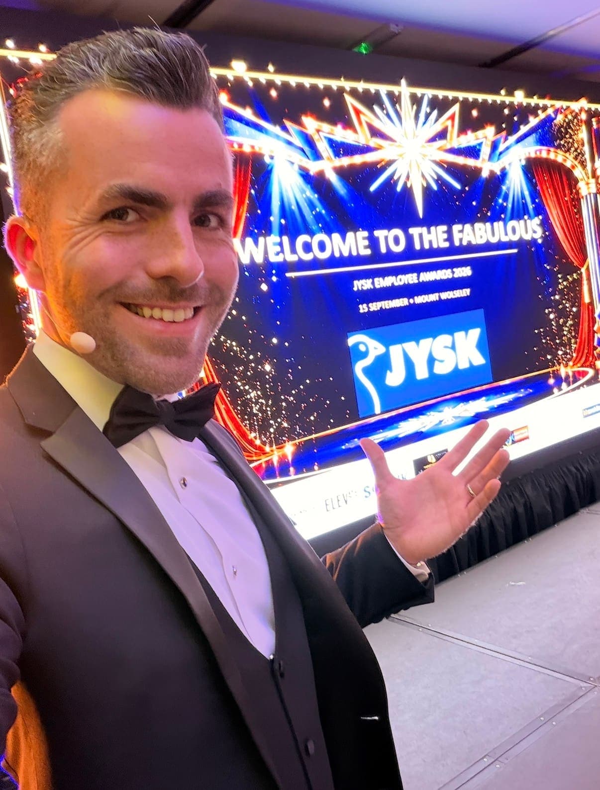 Jamie in a tuxedo and headset microphone smiles in front of a screen that welcomes guests to the JYSK Employee Awards 2026