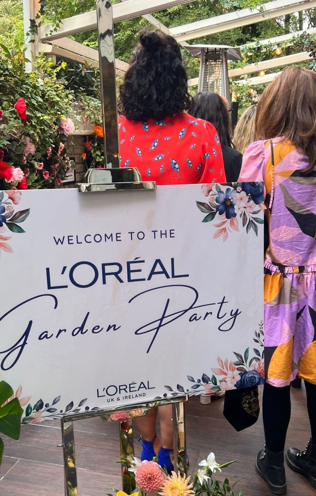 A sign that reads Welcome to the L'Oréal Garden Party, with flowers, guests in summer dresses behind it under a pergola