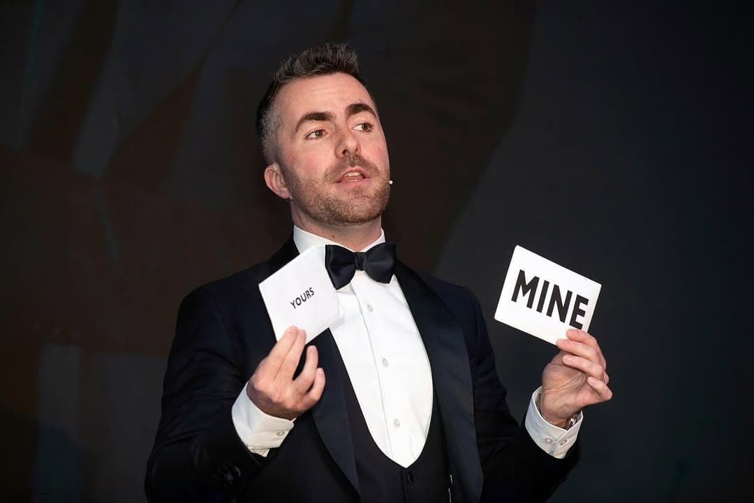 Jamie on stage in a tuxedo with a headset mic, holding up two cards that read YOURS and MINE