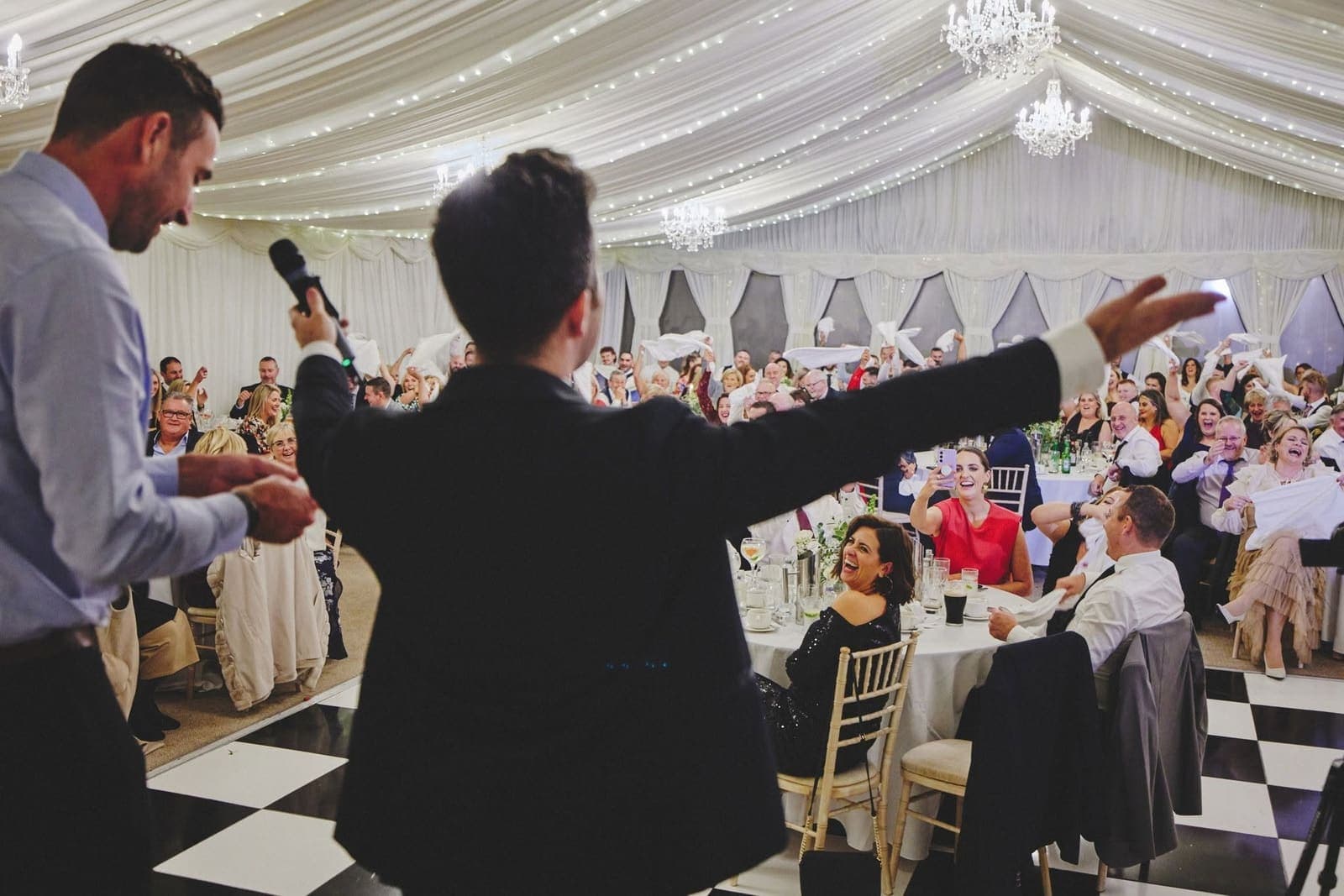 Jamie as MC, microphone in hand, throws his arms out to a full marquee of guests waving napkins