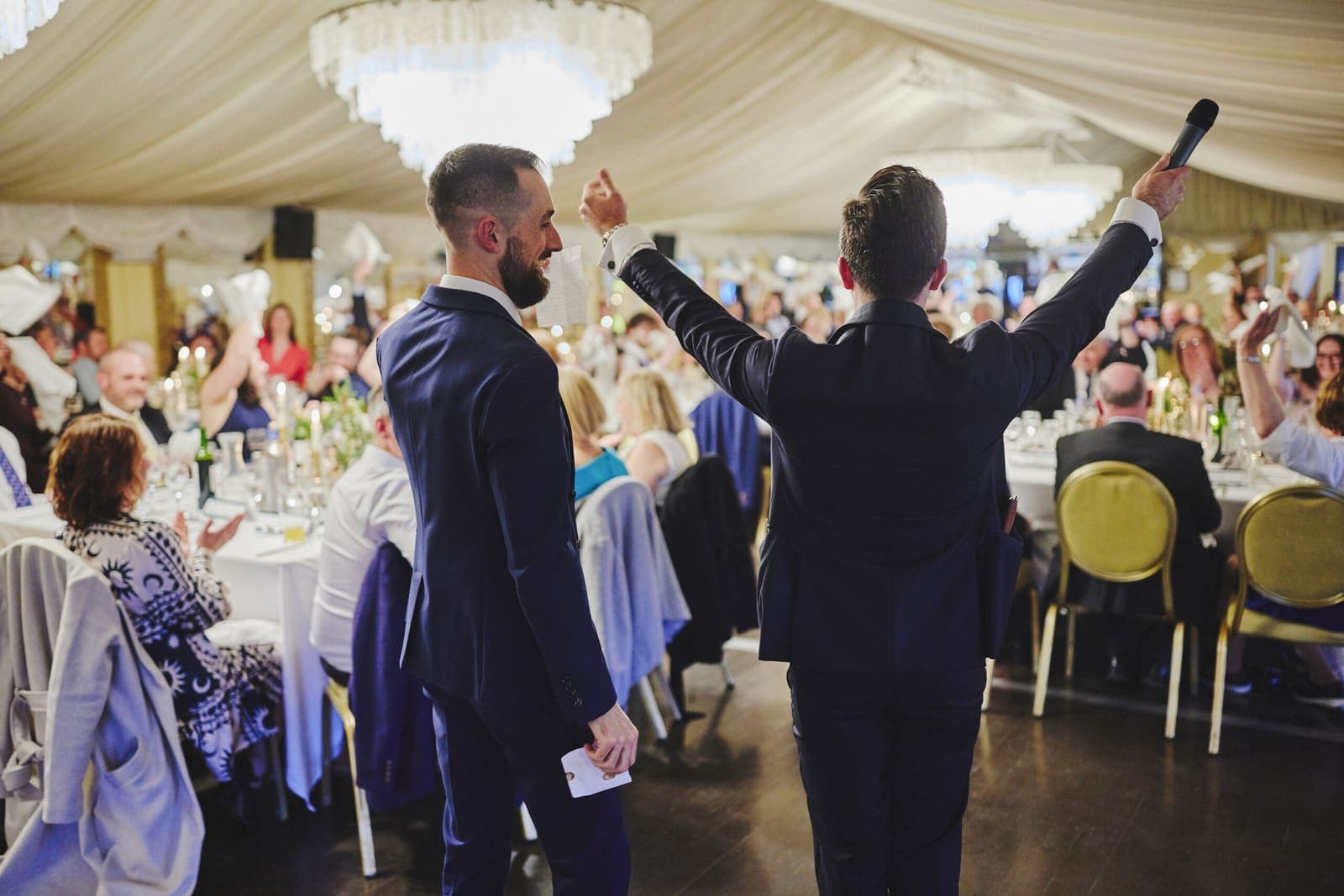 From behind: Jamie and a man raise arms toward a packed wedding marquee with a chandelier