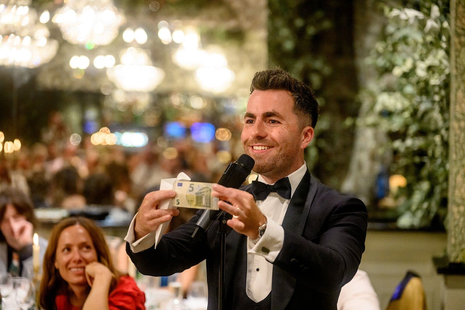 Jamie as MC holds up a banknote with a microphone in a chandelier-lit room; a guest smiles