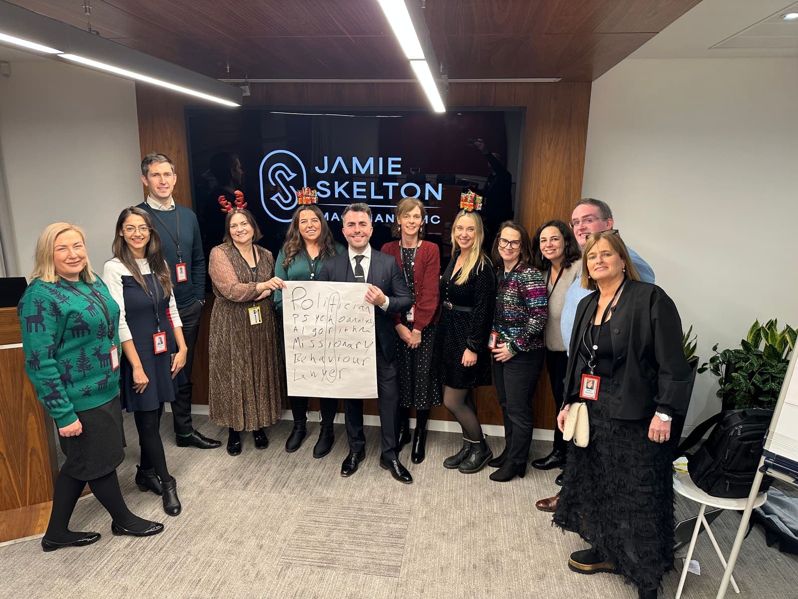 Jamie in a suit stands with office staff with lanyards, some in reindeer and gift headbands, one holding a pad of guessed words, his logo on a screen behind