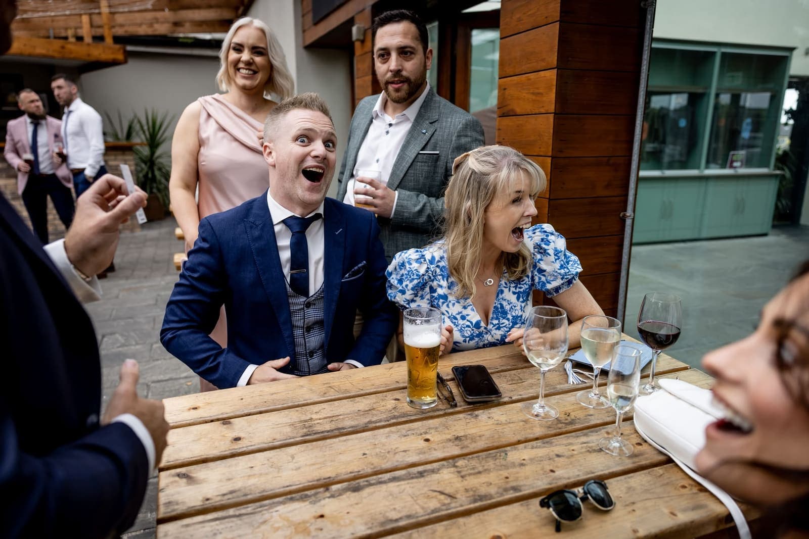 At a relaxed outdoor bench table, a man and a woman stare open-mouthed at a trick while a friend laughs