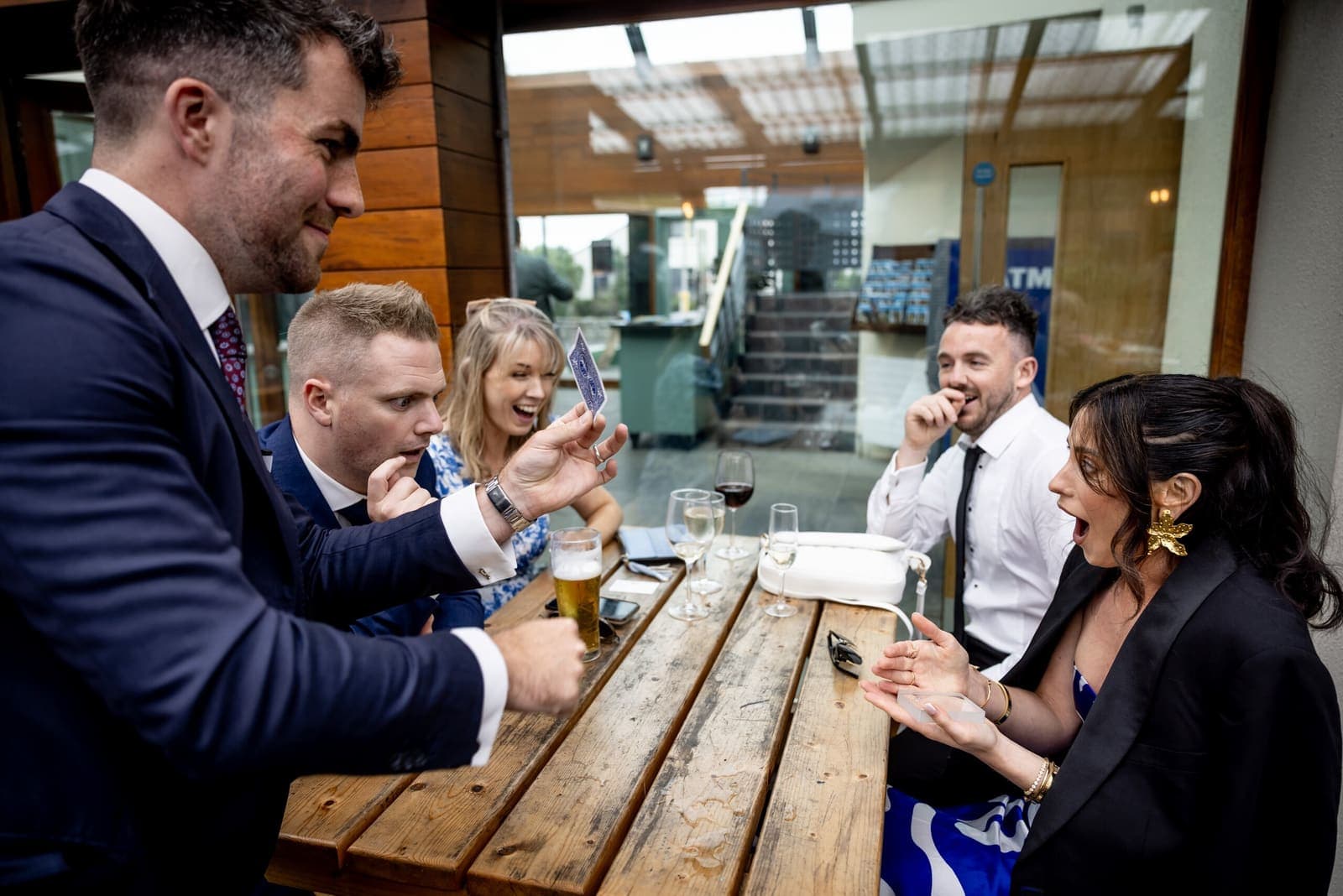 Relaxed guests at an outdoor bench table gasp and laugh as Jamie shows a card
