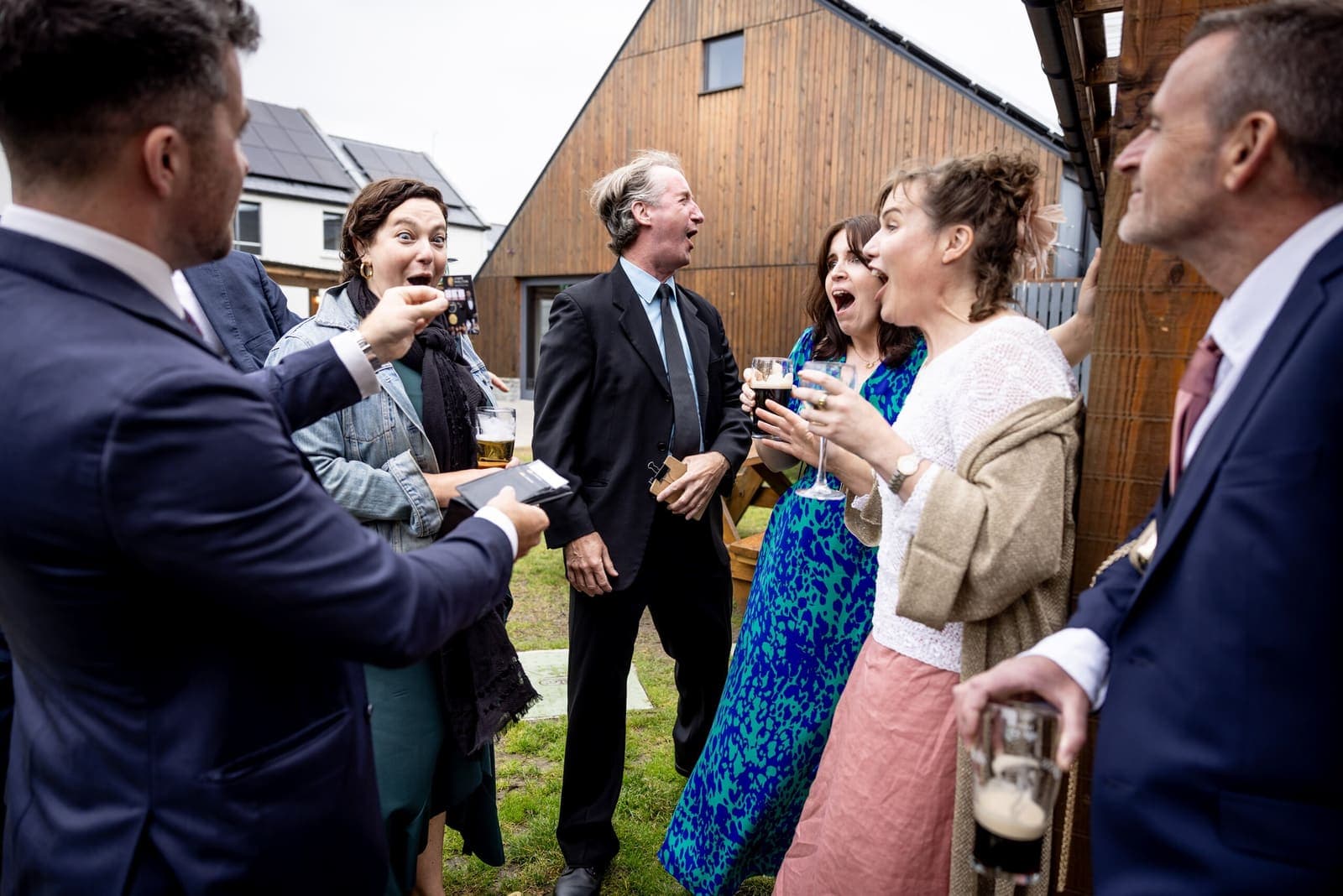 Outdoor wedding drinks reception by a wooden barn; guests react to a trick