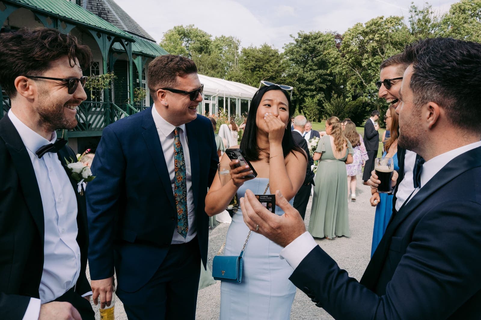 Outdoor wedding drinks; a woman reacts to a trick with three men in suits