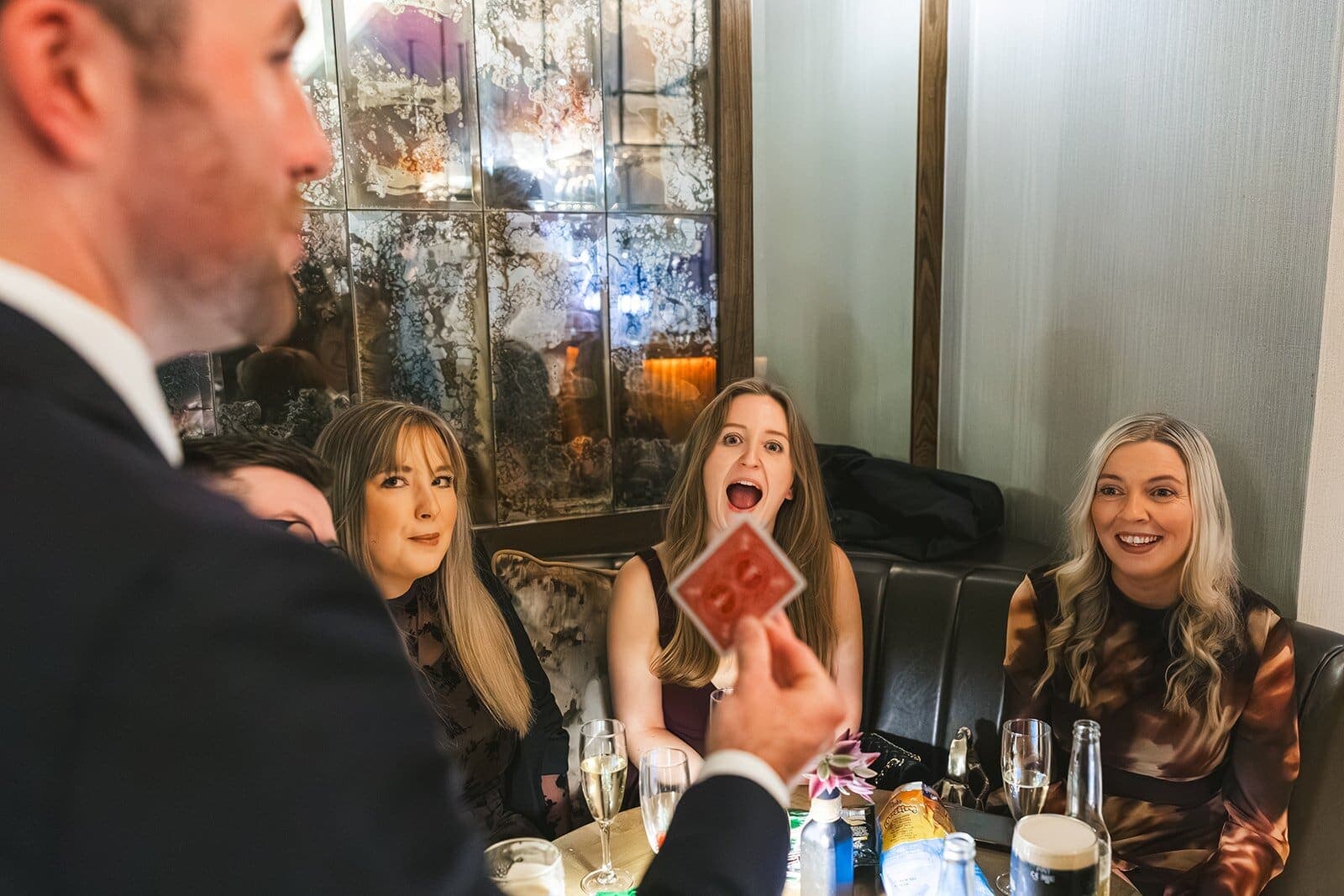 Friends in a restaurant booth gasp and smile as Jamie holds up a card
