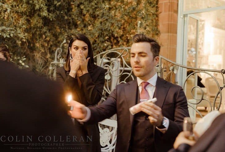 Jamie, in a dark three-piece suit, holds a small flame in one hand and a bottle in the other, while a dark-haired woman behind him covers her mouth with both hands
