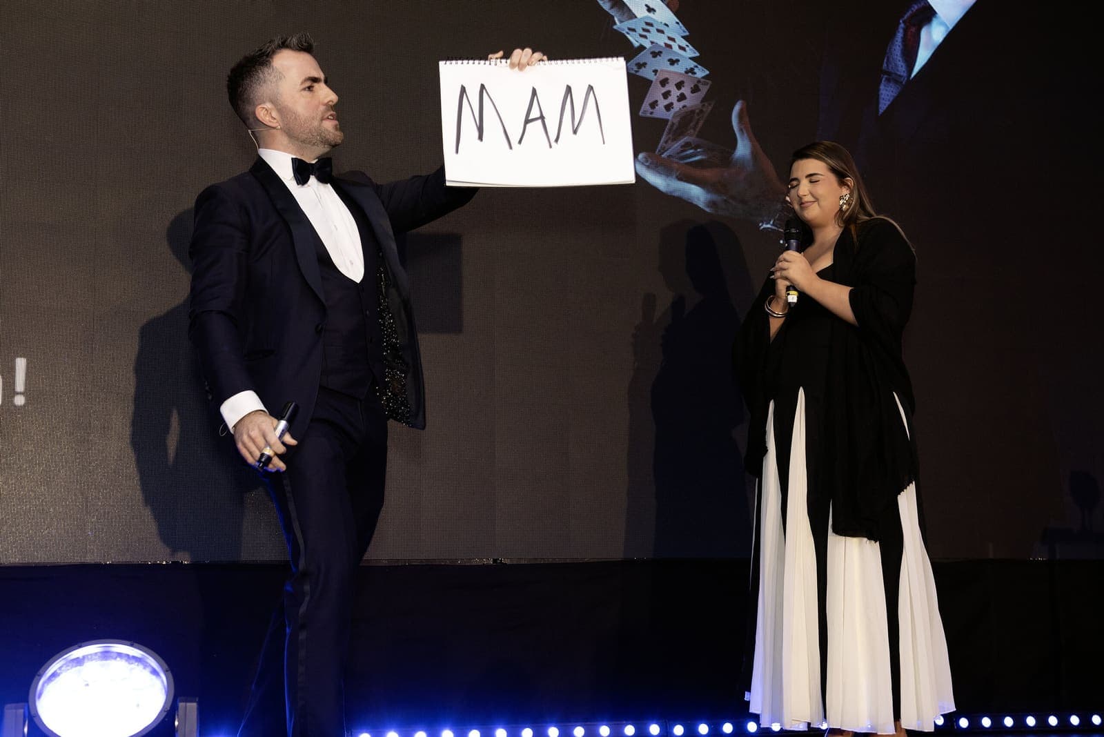 Jamie on stage holds up a prediction card that reads MAM beside a woman with a microphone