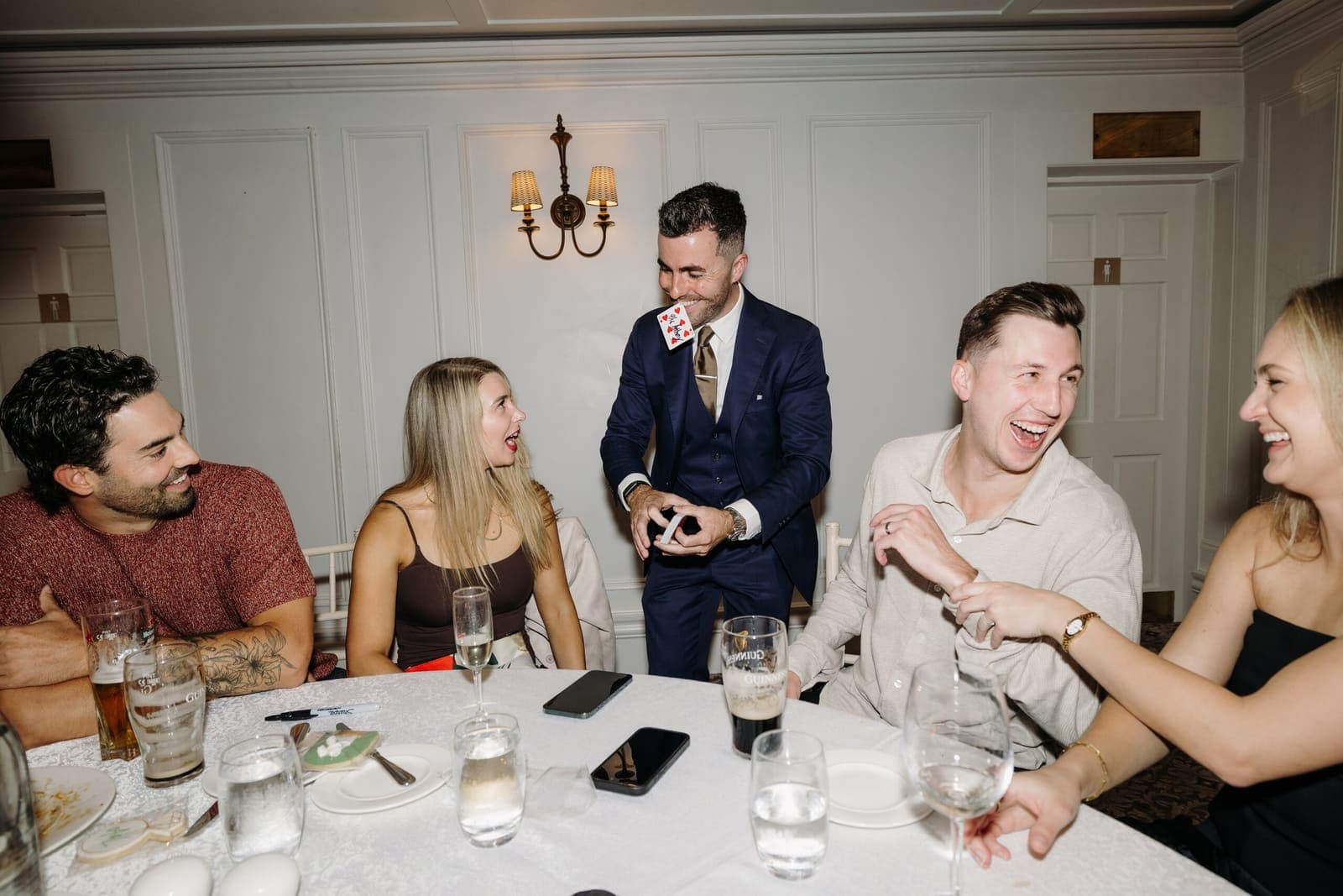 Jamie performs at a round table of laughing guests in a white-panelled room