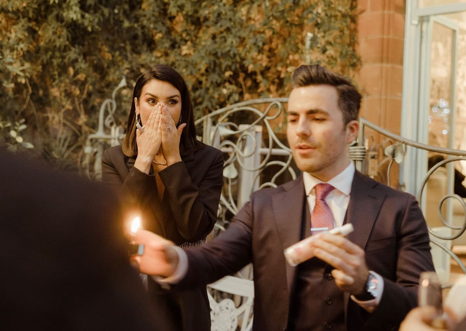 On a terrace, a guest covers her face with both hands as a flame appears from the performer's hand