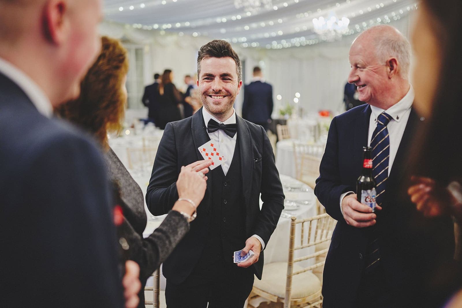 Jamie shows a card to two male wedding guests in a fairy-lit marquee