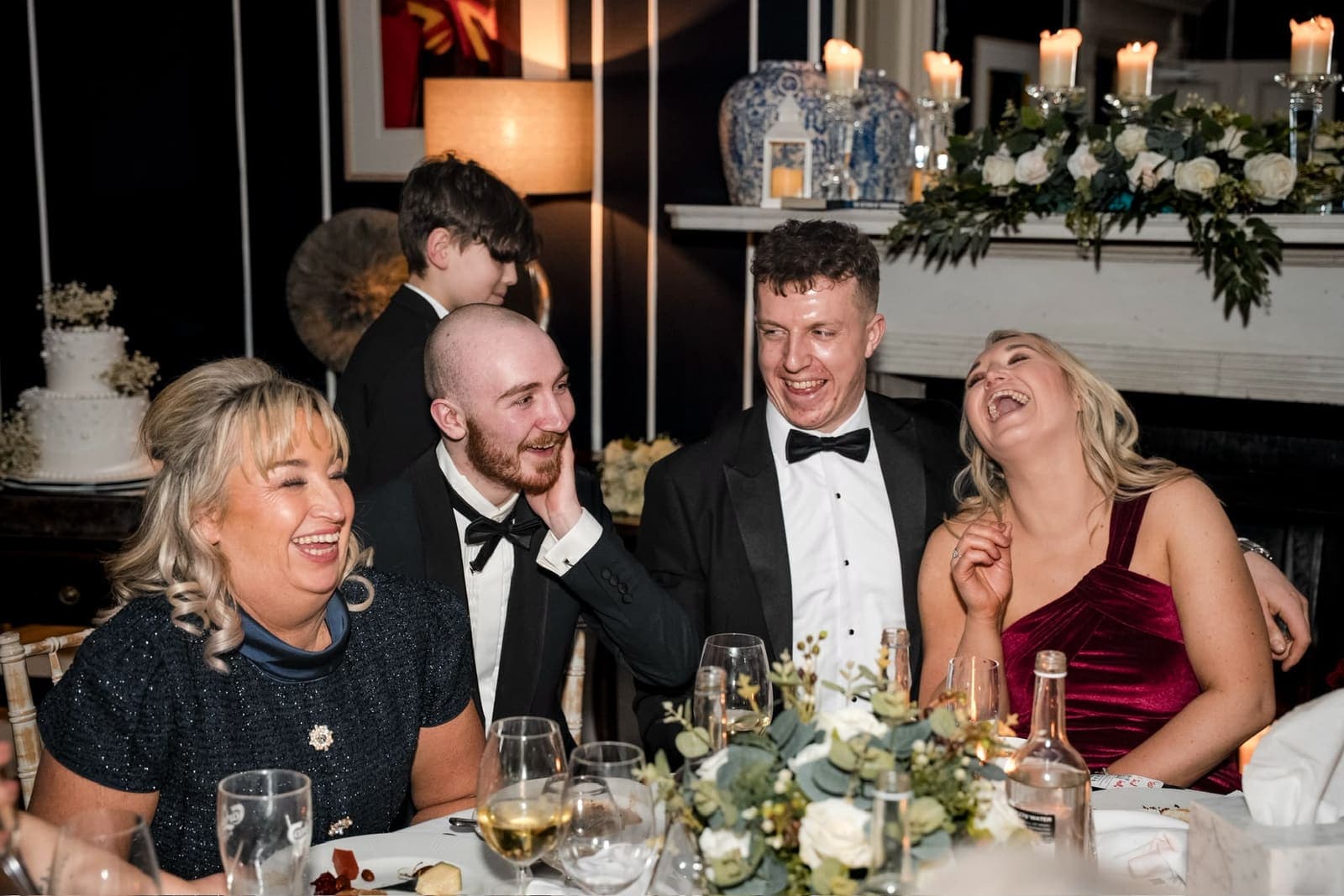 After dinner, four guests at a round table in black tie and dresses laugh hard, one with her head thrown back