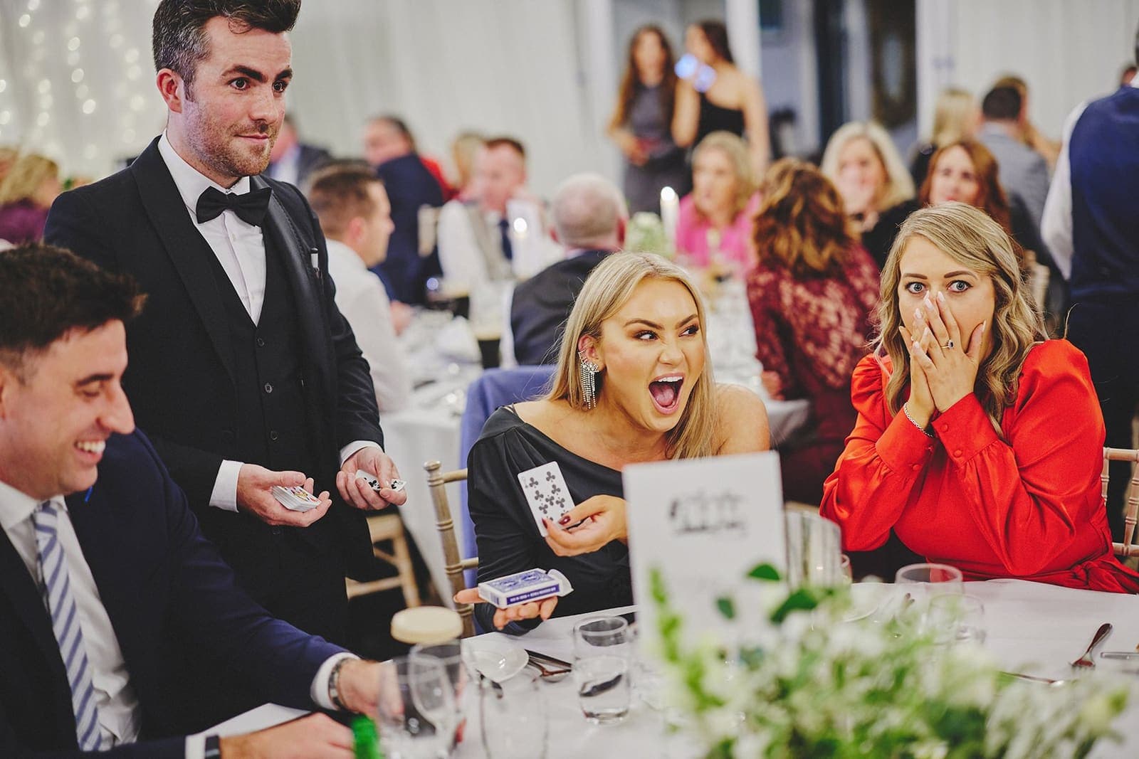 At a wedding dinner table, a guest in red covers her face with both hands while the woman beside her gasps at a card