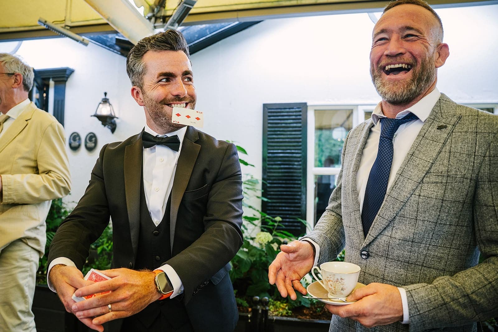 At a drinks reception, Jamie holds a card in his teeth and a guest with a teacup laughs