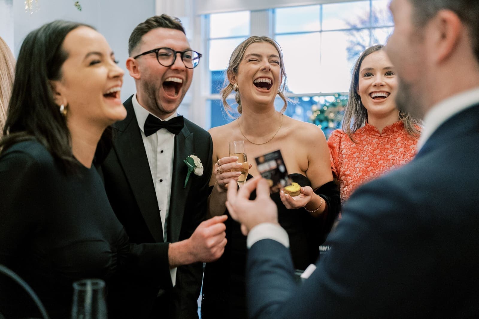 Wedding guests in black tie and dresses react with open-mouthed delight to a card trick