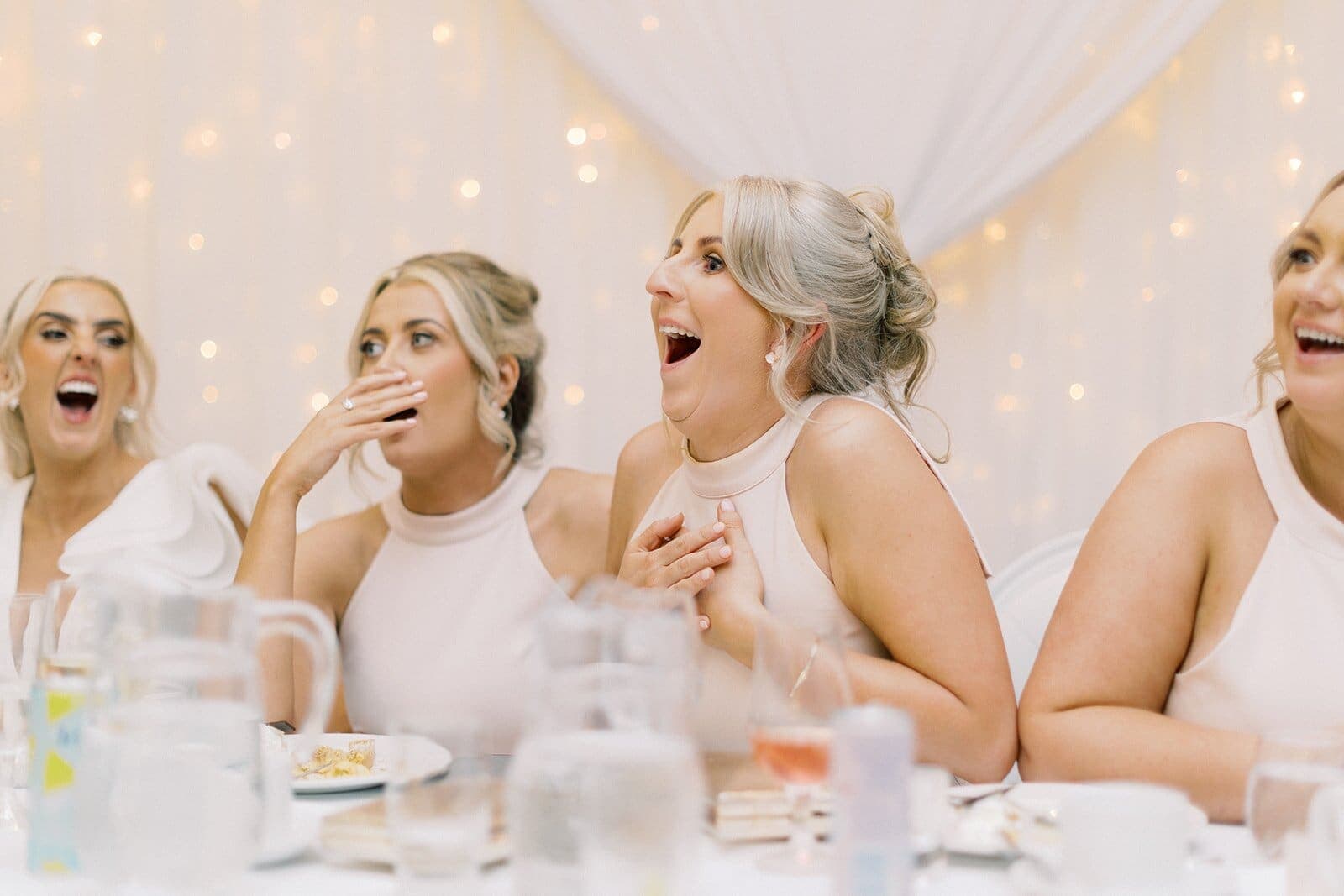 Guests in white dresses gasp, hands over mouths, in front of fairy lights