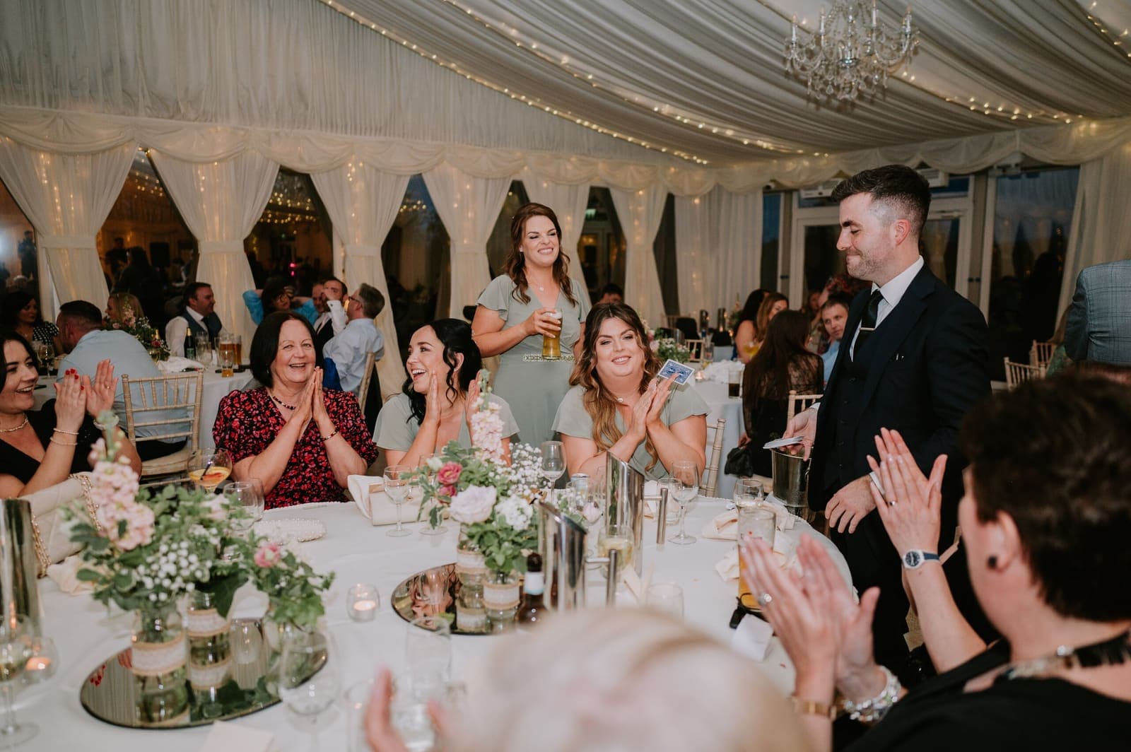 Wedding guests at floral tables applaud in a fairy-lit marquee