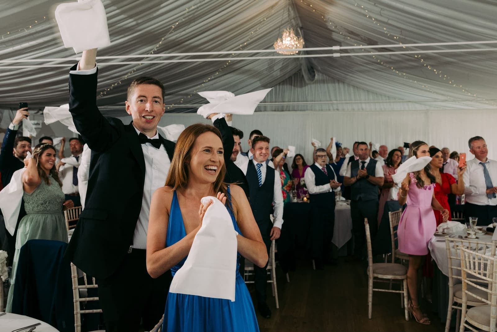 A full wedding marquee waves white napkins in the air, guests laughing and cheering