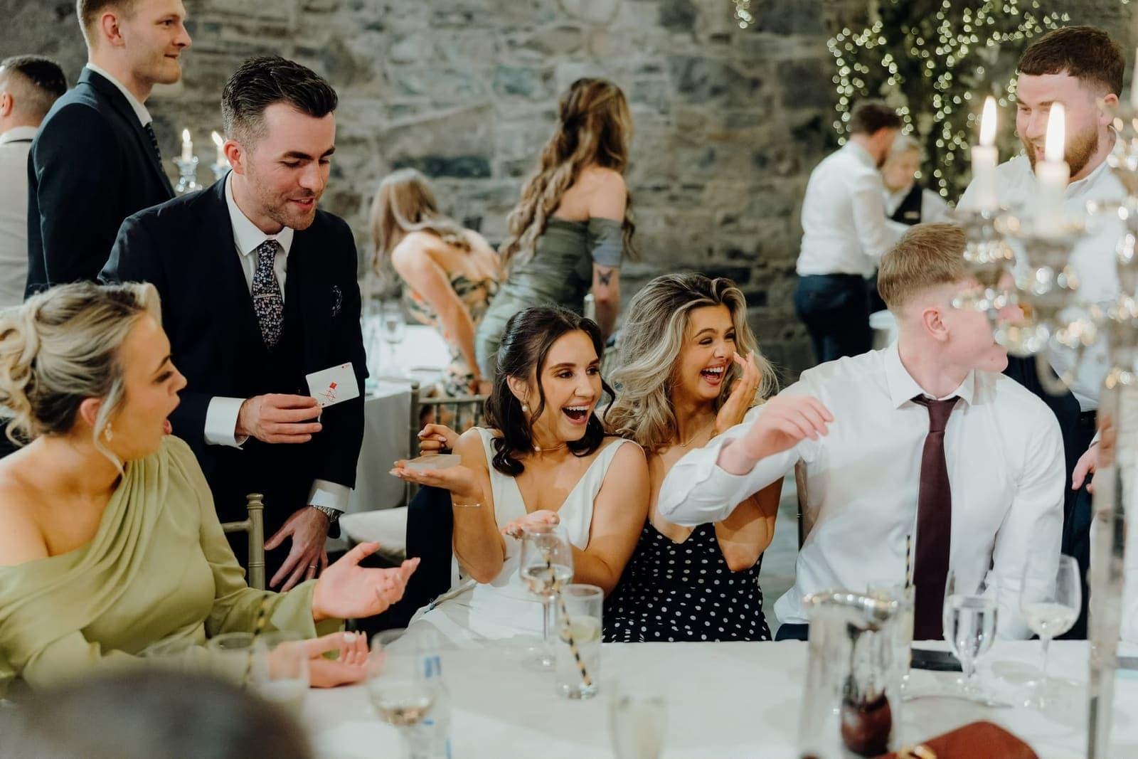 Guests at a table in a stone hall cover their mouths and laugh at a card trick