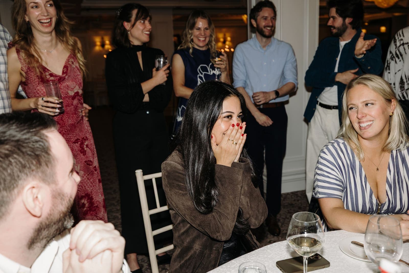 After dinner, a guest at a round table covers her mouth in shock while the guests around her laugh