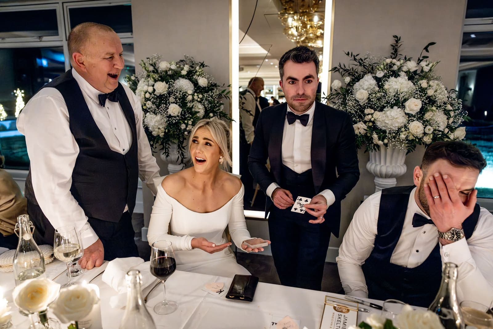 At the top table, the bride laughs open-mouthed and a man beside her hides his face in his hand as Jamie holds up a card