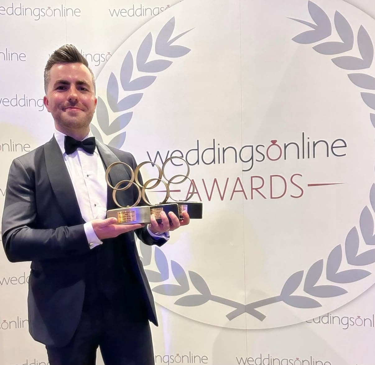 Jamie in a tuxedo holds three weddingsonline awards in front of the weddingsonline Awards backdrop