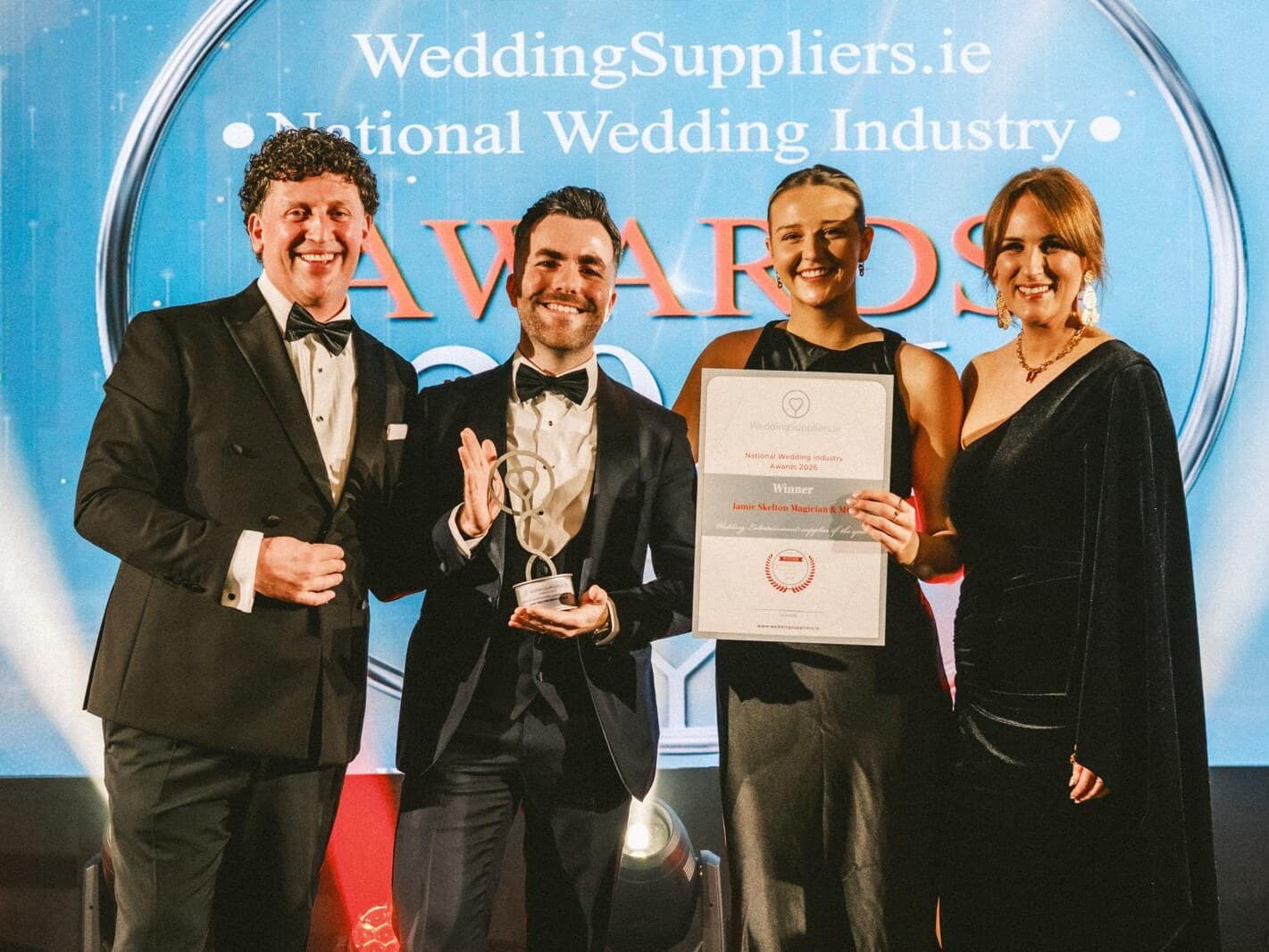 Jamie in black tie holds his Weddingsuppliers.ie trophy on stage beside a man in black tie and two women, one holding his Winner certificate, under the National Wedding Industry Awards screen