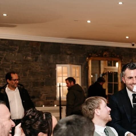 Jamie holds up a card to a table of smiling guests at Kelly and Owen's wedding at Tankardstown House