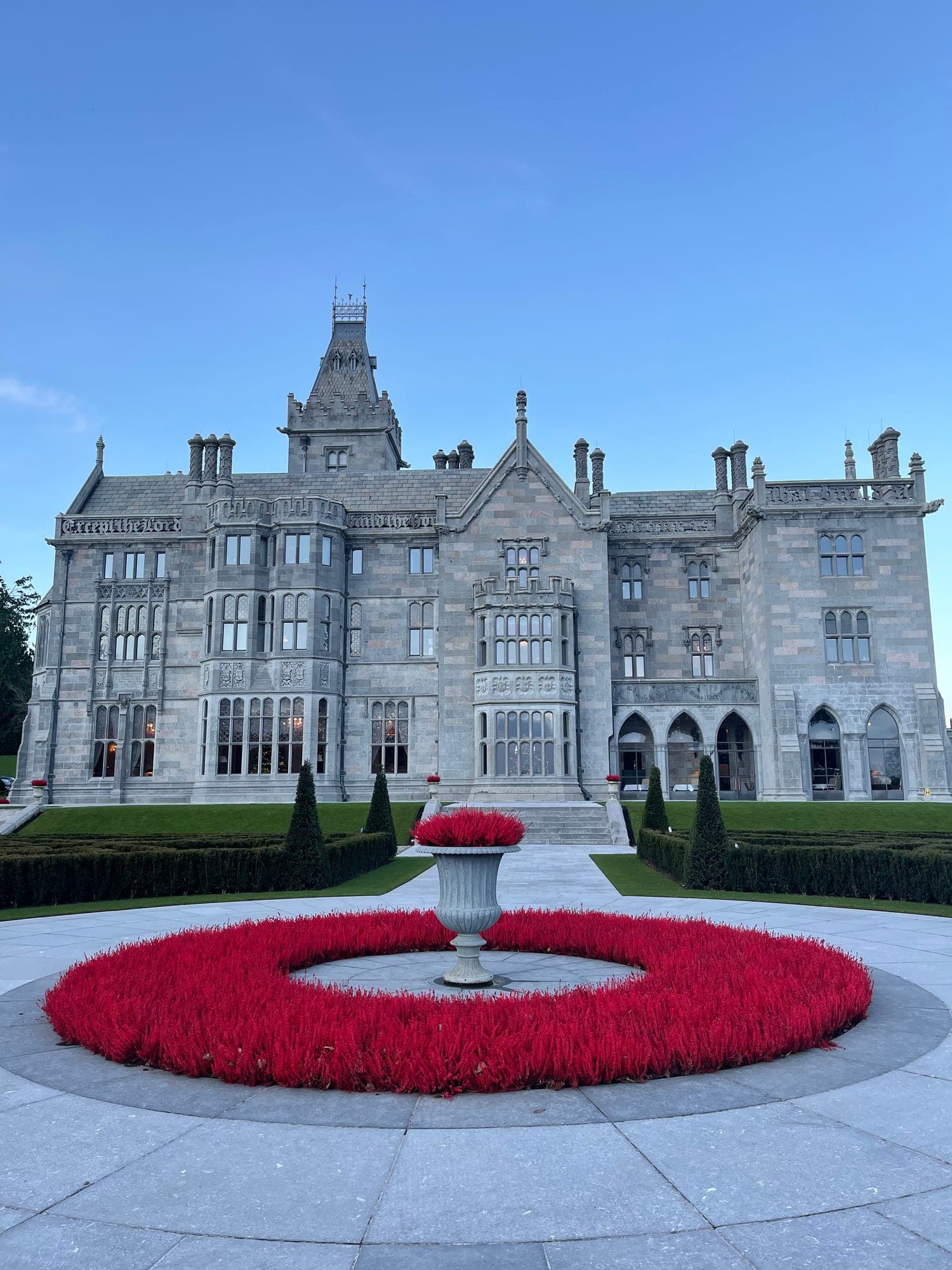 The stone garden front of Adare Manor under a blue sky, behind a round bed of red flowers and an urn