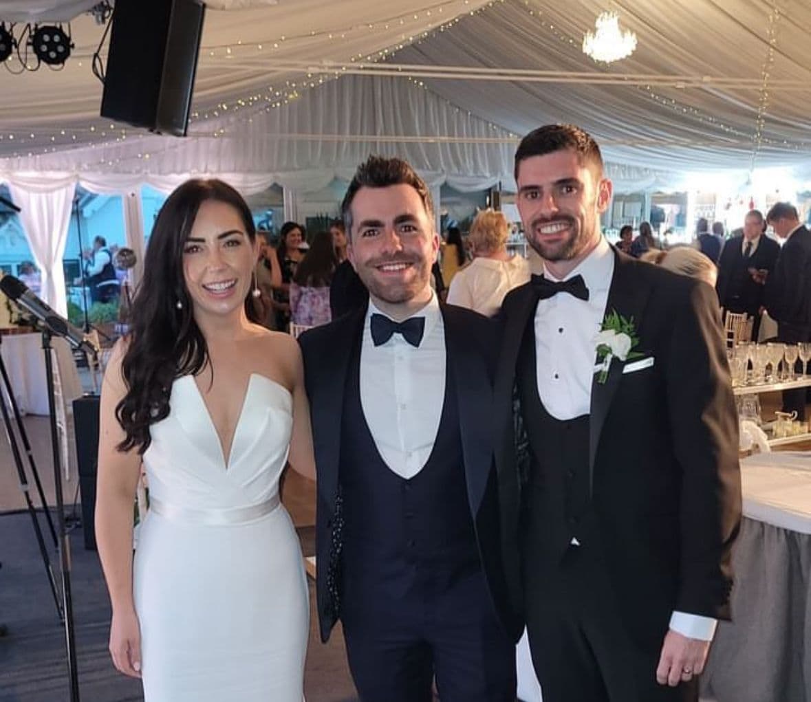 Laura and Darragh smile beside Jamie in black tie, under fairy lights in a marquee