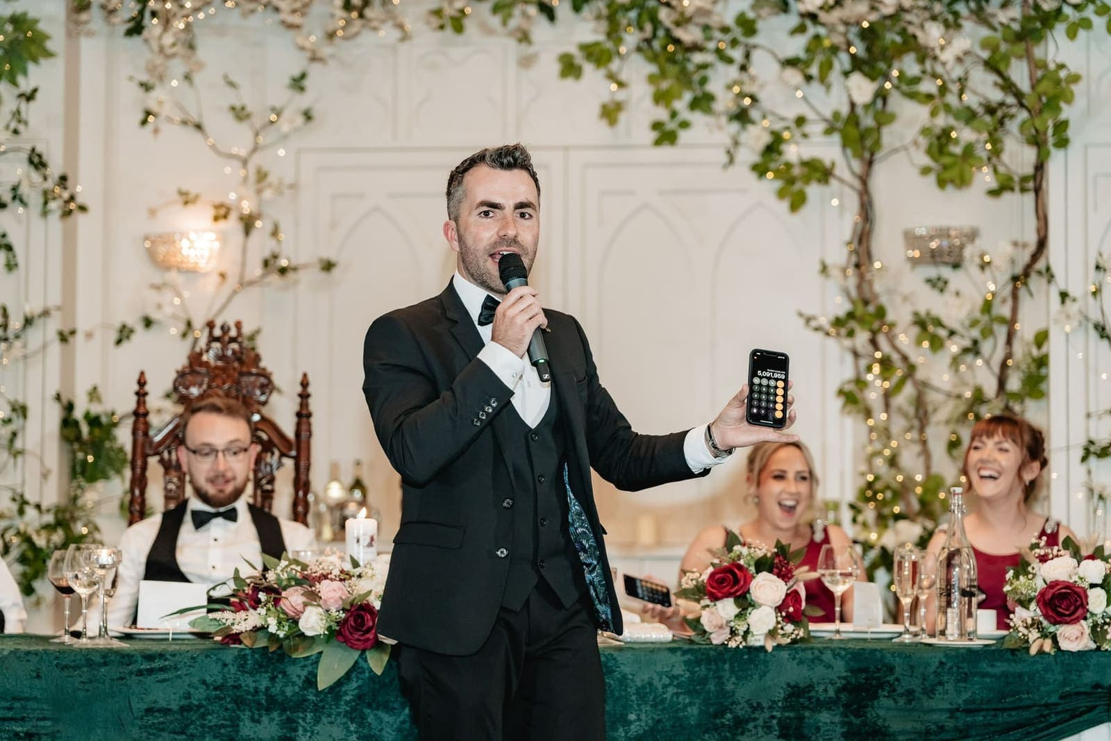 On the mic at Alex and Suzanne's top table, Jamie holds up a phone calculator as two bridesmaids in red laugh out loud