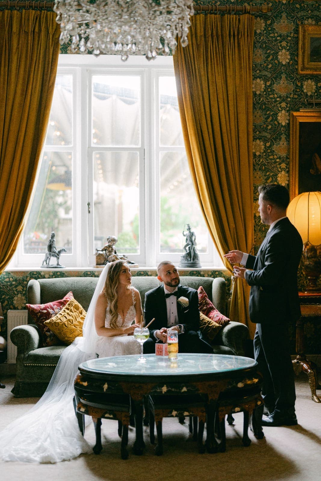 Under a crystal chandelier, Amy and Luke look up at Jamie from a sofa by a tall window
