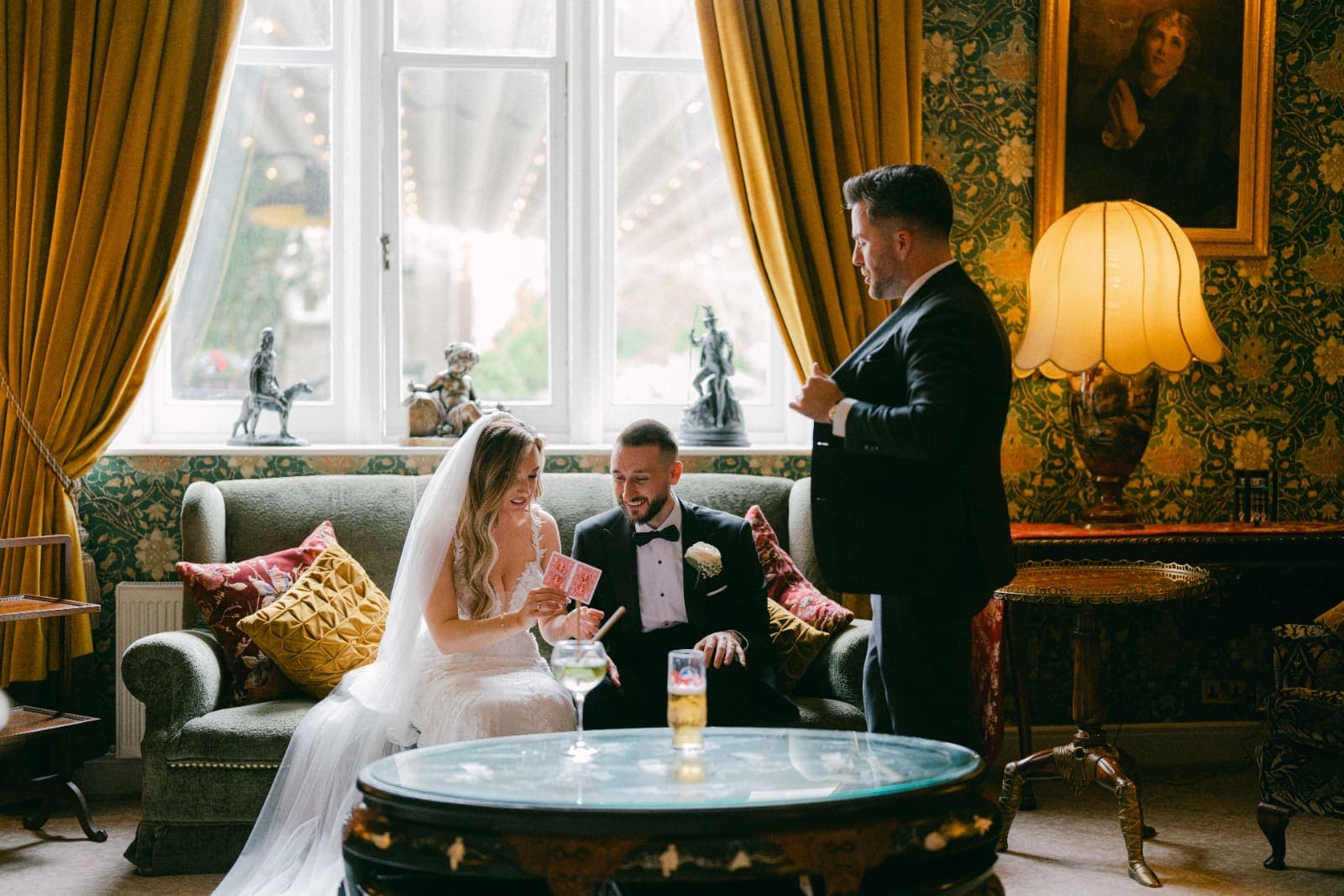 In a room with green wallpaper and gold curtains, Amy holds up two cards as Luke smiles and Jamie stands beside them