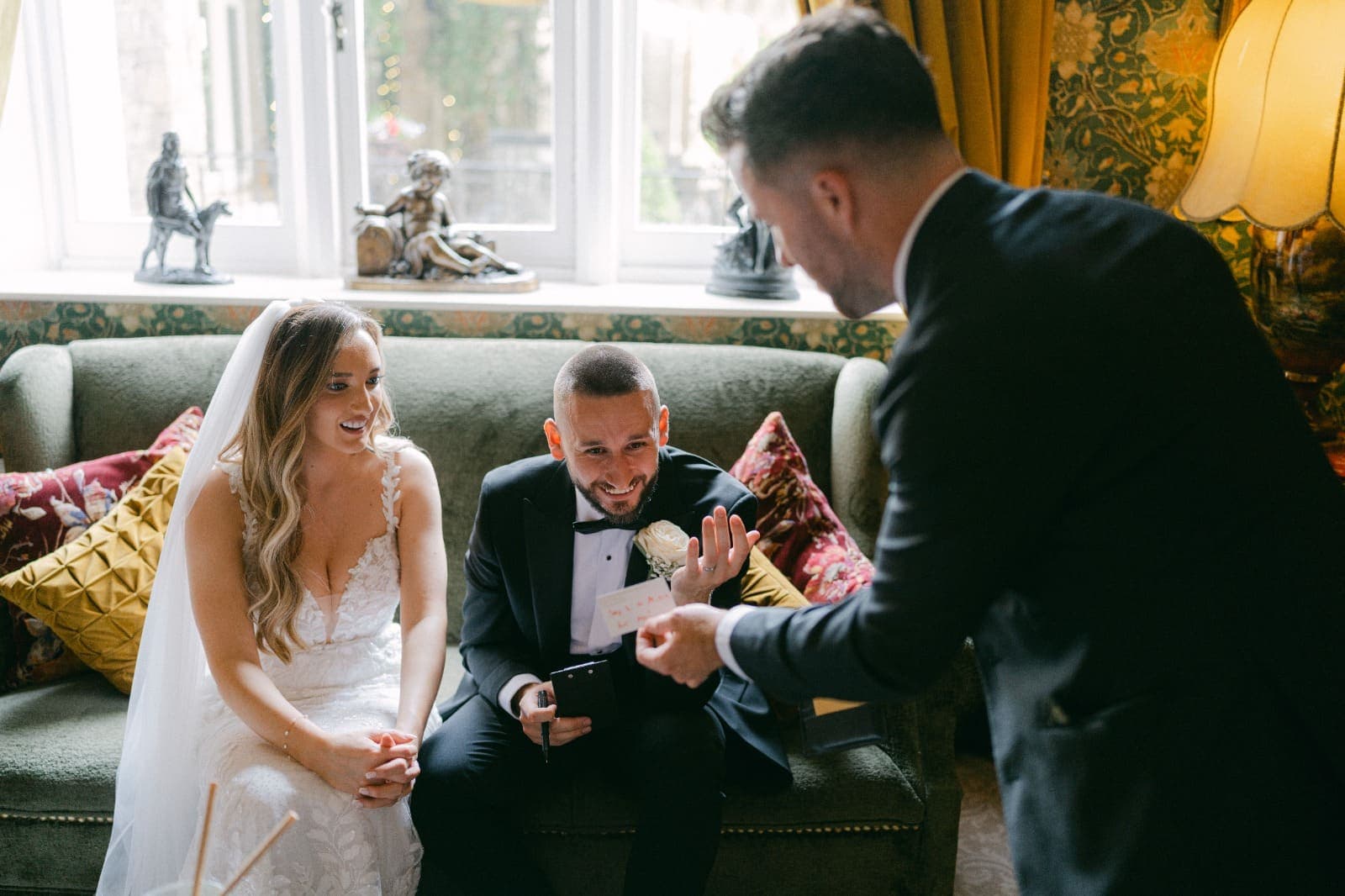 On a green sofa at Cabra Castle, Luke grins and throws up a hand as Jamie shows him a small card, and Amy smiles