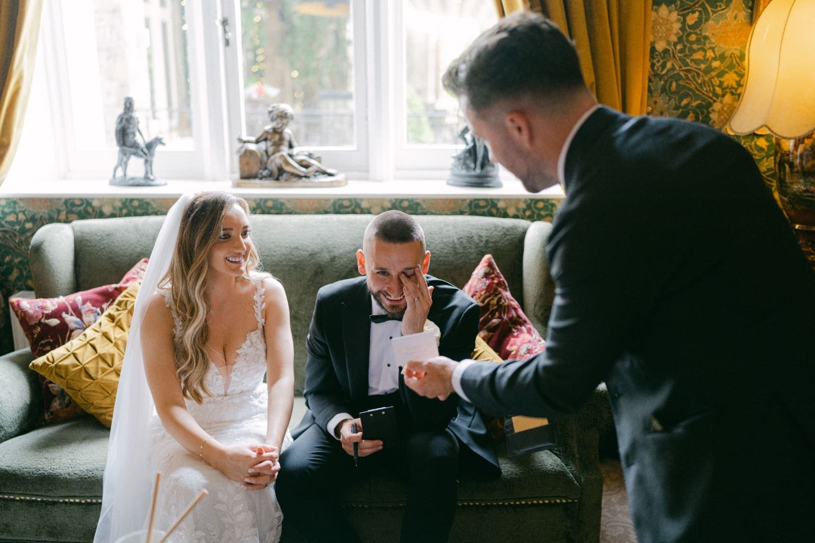 Luke laughs and wipes his eye as Jamie holds out a small card, and Amy smiles beside him on the sofa