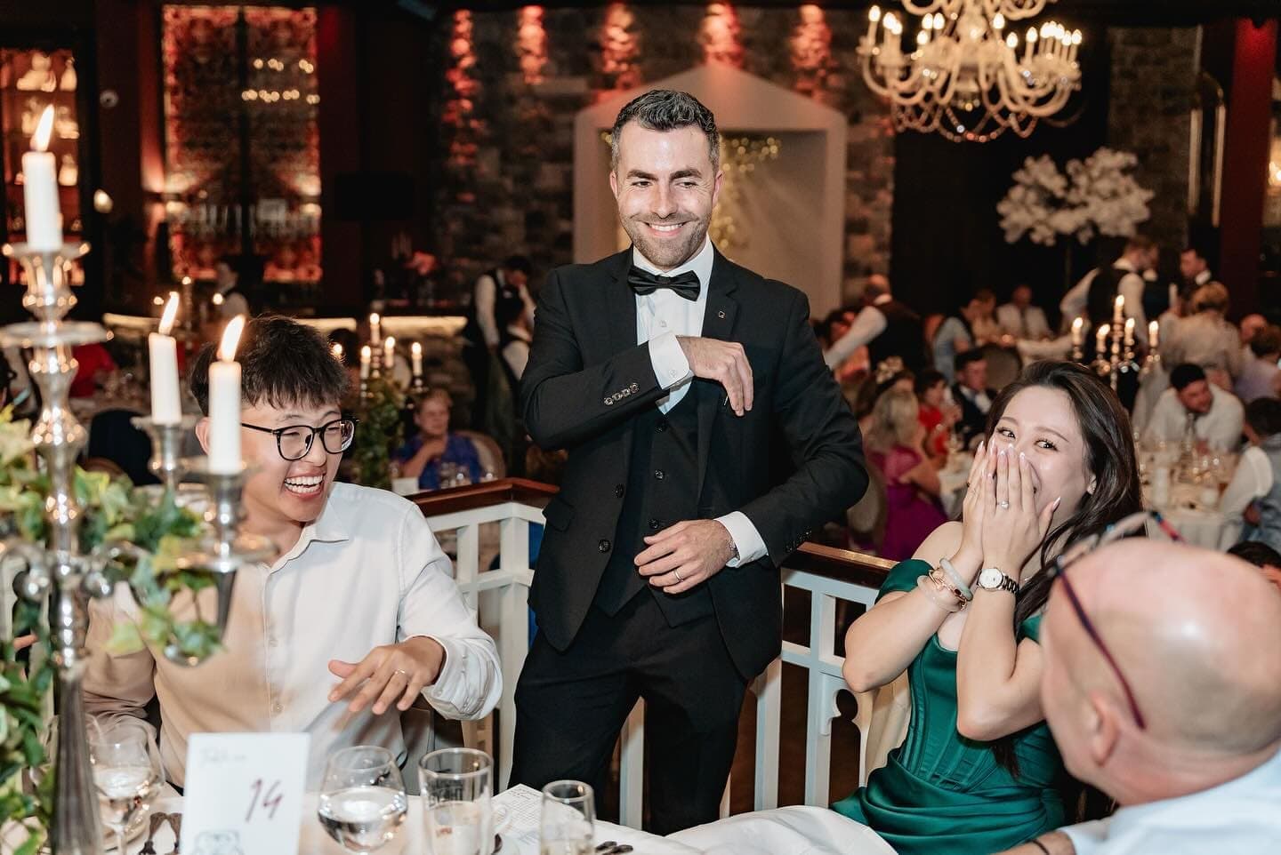 A guest in green covers her mouth and the man beside her laughs as Jamie smiles at their table